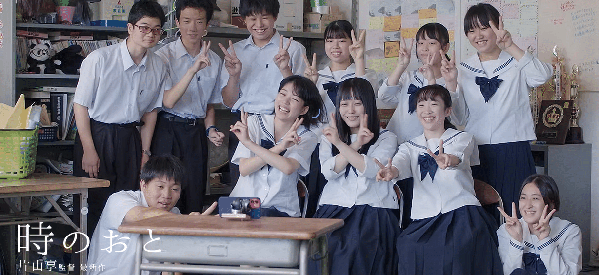 高校生たちが部室で集合写真を撮っている。机にスマホを立てて、女子生徒6人と男子生徒4人が、笑顔でピースサインをしている