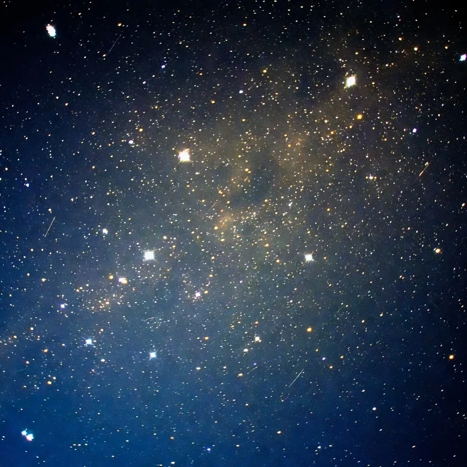 深い青と黒の背景に、大小さまざまな無数の星が散りばめられた星空の写真。十字状の光筋を持つ特に明るい星が数個点在し、中央から右上にかけて密度の高い黄金色の星々や淡いモヤのような星雲が広がっている。