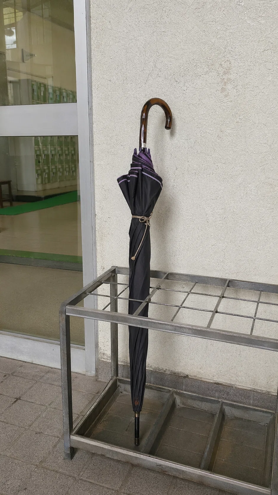 体育館入口付近の金属製傘立てを写した事務的な記録写真。ほかに傘はなく、黒地に鈍い紫の縁取りがある古い長傘が一本だけ立てられている。べっこう色の曲がり手が見え、施設職員が記録用に撮影したような乾いた構図になっている。