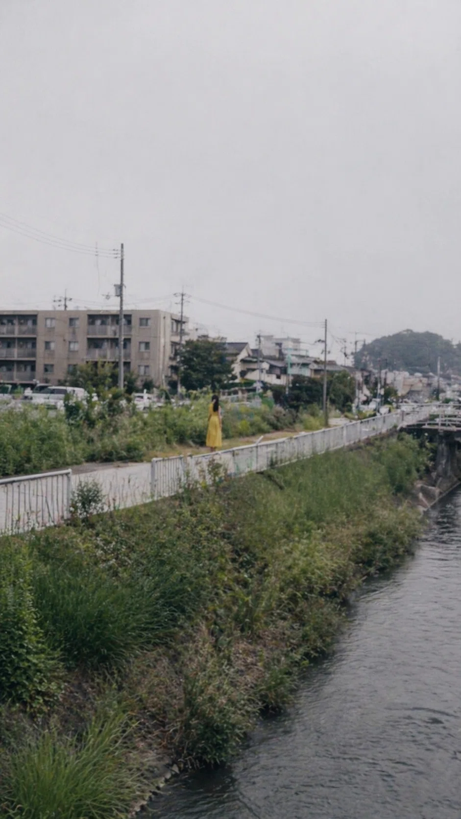 川沿いの遊歩道を少し離れた位置から撮影した写真。曇天の河川敷風景の奥に、黄色い衣服を着た細身の高い女性が一人だけ立っている。距離があるため顔立ちは判別できず、周囲の景色だけが妙に鮮明に見える。