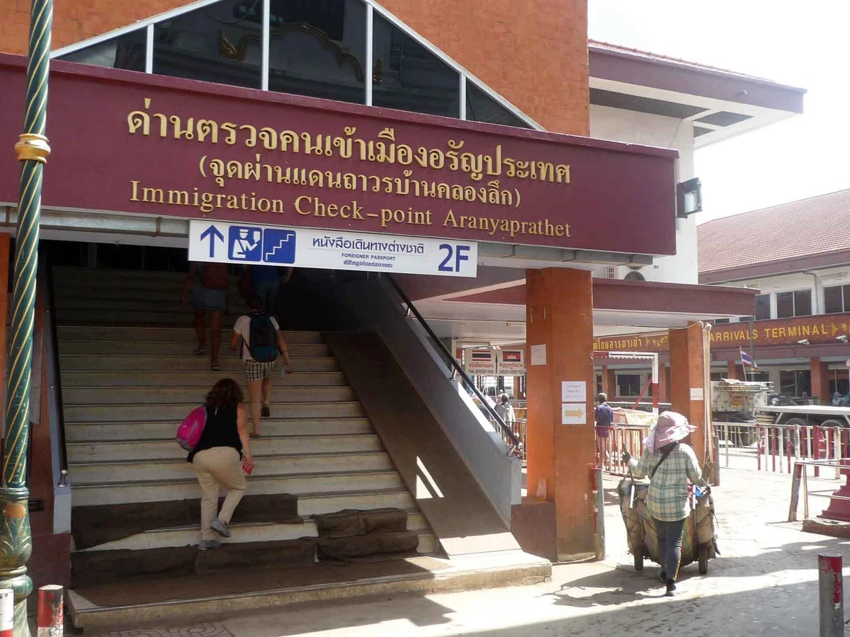 “Immigration Check-point Aranyaprathet” と書かれた建物の入口。階段を旅行者が上がり、右側には荷物を運ぶ人がいる。