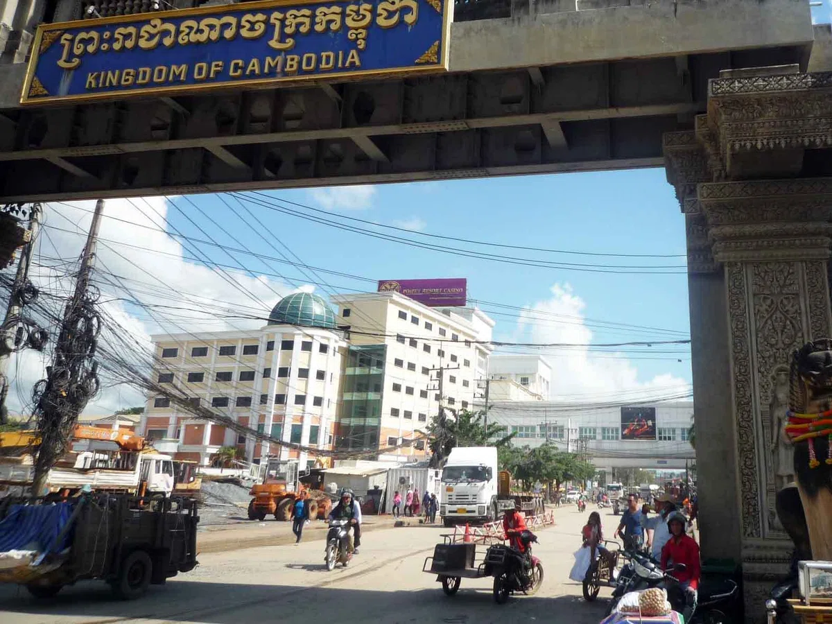 “KINGDOM OF CAMBODIA” と書かれた門の先に、カジノの建物が見える。道路ではトラックやバイク、人々が行き交っている。