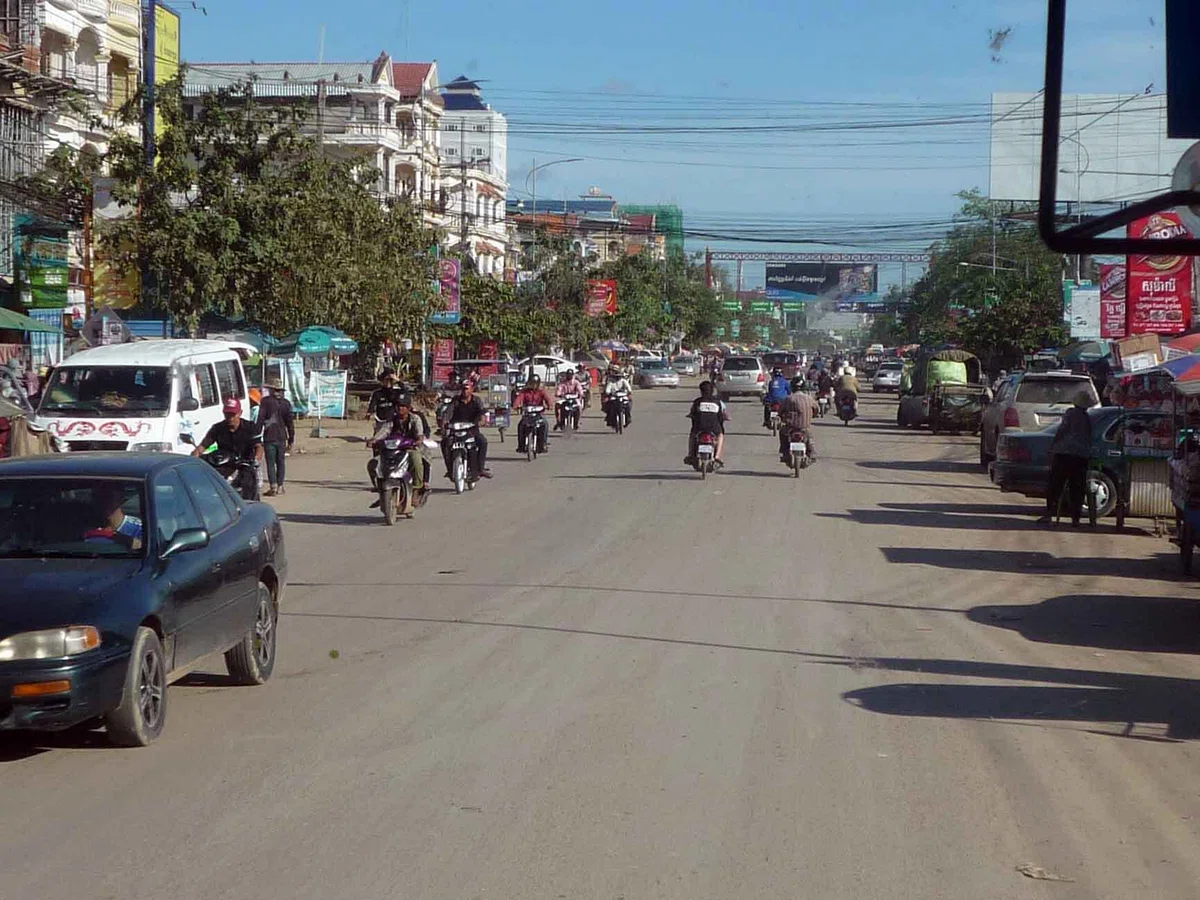 バスの車窓から見たカンボジア側の道路。車やバイクが走り、通り沿いに建物や看板が並んでいる。