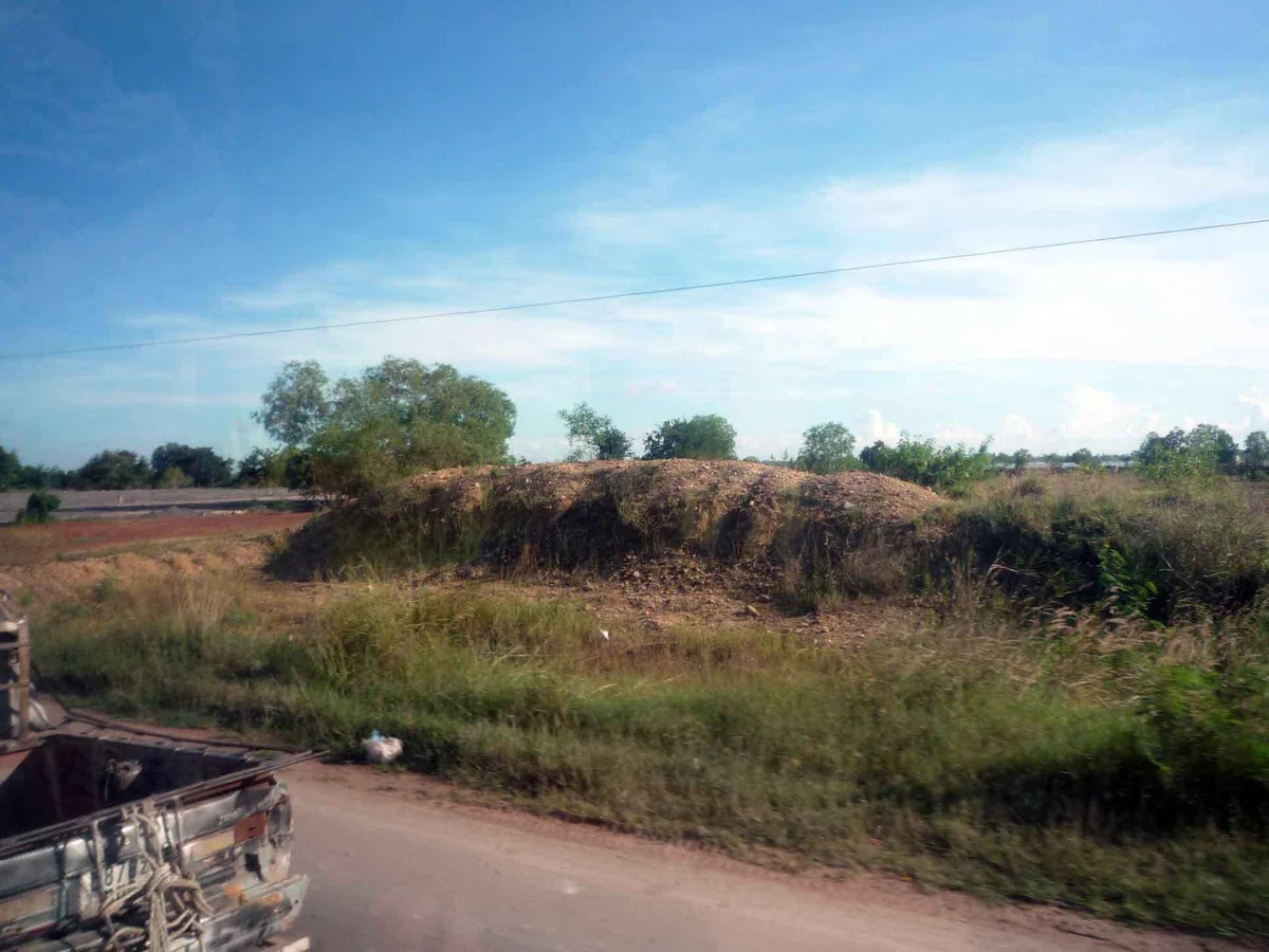 バスの車窓から見たカンボジアの郊外の風景。道路脇に草地と赤土の地面が広がっている。