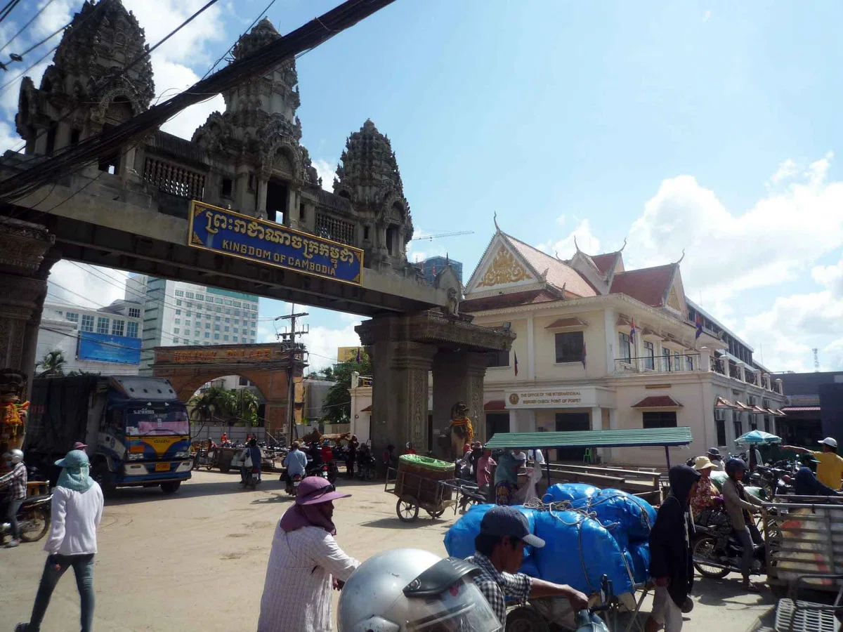 「KINGDOM OF CAMBODIA」と書かれた国境ゲートの前を、バスやバイク、荷車、人々が行き交っている。