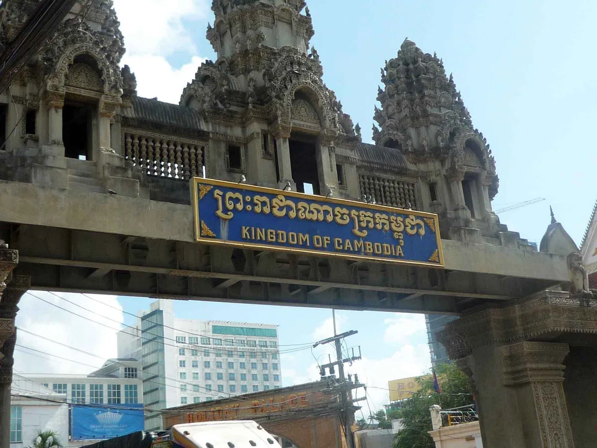 “KINGDOM OF CAMBODIA” と書かれたカンボジア側の国境ゲート。門の下に建物や車両が見えている。