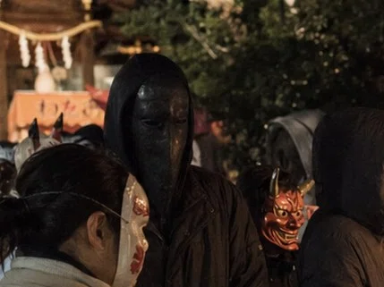 夜の祭礼会場を近い距離から撮影した写真。面をつけた複数の参加者に囲まれる中、中央に黒く細長い面をつけた人物が立っている。周囲の面は狐や鬼など祭礼用の意匠だが、中央の人物だけは口のない黒い面をつけており、質感も異なる。背後には屋台の灯りと神社境内の装飾が見える。