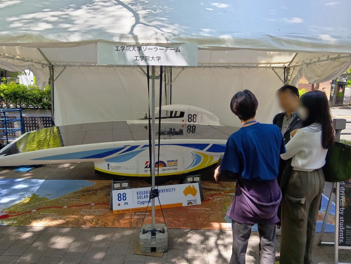 地元大学の学生がフォーミュラカーについて説明し、来場者が車両に乗り込みながら聞いている