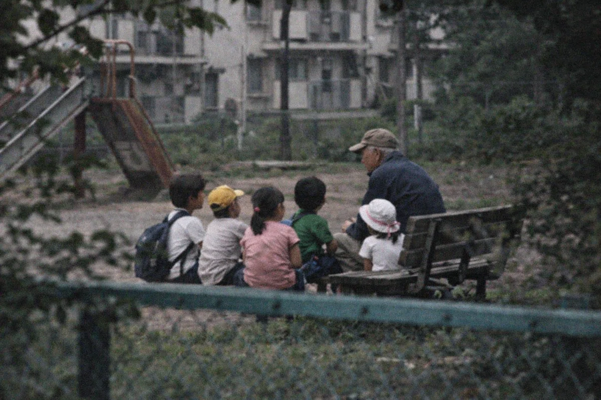 団地内の公園を少し離れた位置から撮影した記録写真。ベンチの周辺に数人の児童が座っており、その前に帽子をかぶった高齢男性がいる。背景には古い団地建物、遊具、植え込み、柵などが見える。男性の顔は距離と角度のため判然とせず、普通の地域写真のように見える。
