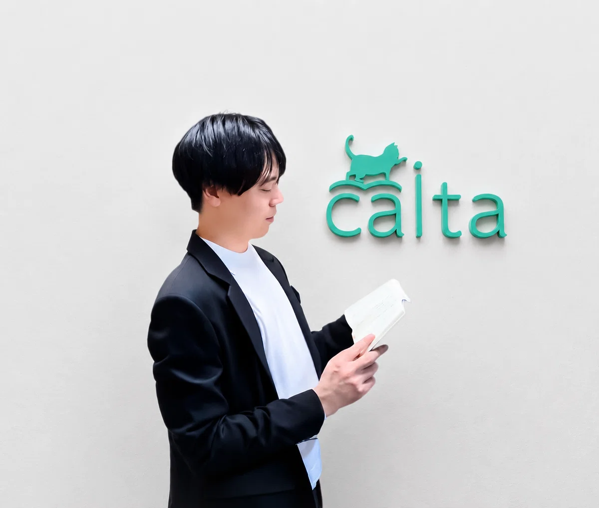 本を手に取る柳村代表。黒いジャケットの下に白いシャツを着ており、壁には緑色のCaitaのロゴが描かれている。