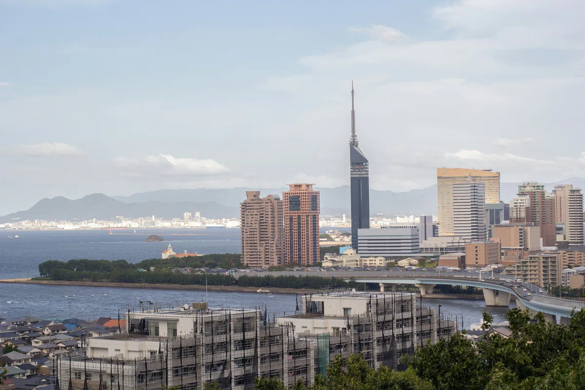 海沿いの都市風景を写した横長の写真。画面上部の大部分に淡い青空と白い雲が広がっている。中央右寄りには、細長いアンテナ状の先端を持つ高い塔状の建物があり、その周囲に高層マンションやオフィスビルが複数並んでいる。建物群の背後には、海を挟んで山並みと市街地が見える。画面左側には広い海面があり、海上に小さな島や船が見える。中央下には樹木が連なる海沿いの緑地があり、その右側から橋が伸びている。手前には足場や養生ネットが設置された建物、住宅の屋根、右下に樹木の葉が写っている。