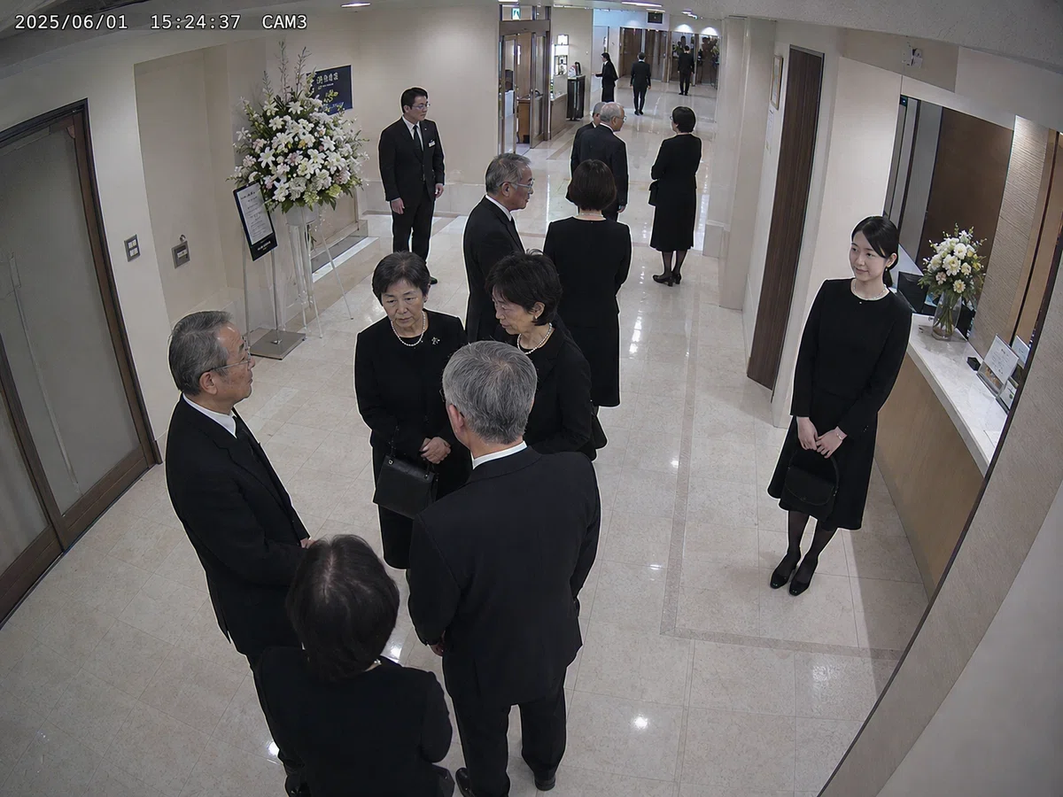 葬儀場のロビーを上方から撮影した監視カメラ映像。黒い喪服の参列者たちが中央で会話しており、奥には式場出入口と受付周辺が見える。画面右側の受付脇に、黒い喪服を着た若い女性が一人で立っている。女性は髪をまとめ、小さなバッグを持ち、周囲の参列者とは少し離れている。画面左上には日時とカメラ番号が表示され、防犯カメラの記録映像に見える。