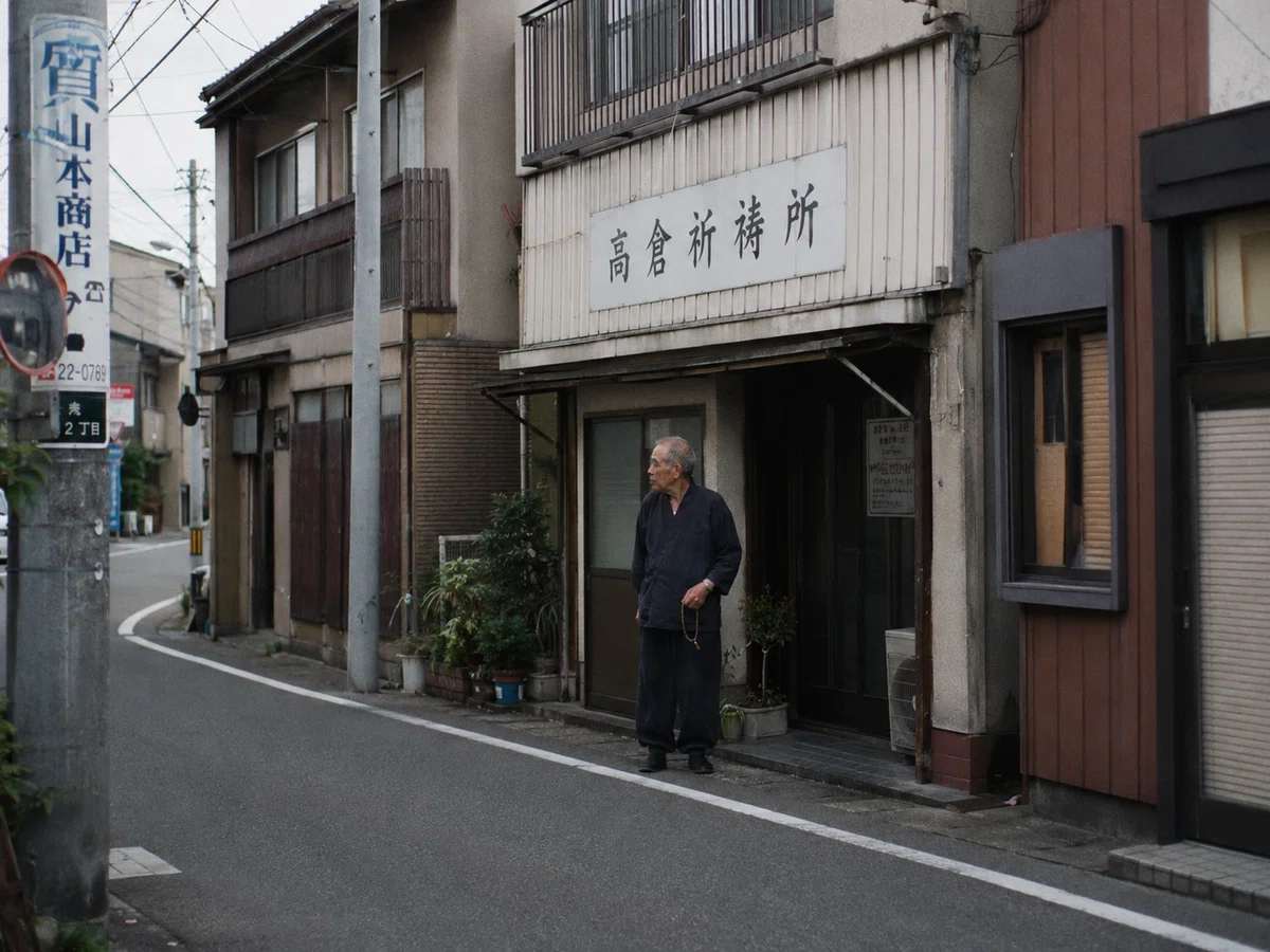 水尾町二丁目の古い通りにある、高倉祈祷所の前を道路側から撮影した記録写真。古い建物の一階部分に大きな看板が掲げられ、入口付近に七十代の男性が一人で立っている。男性は濃色の作務衣風の服装で、手には数珠を持っている。周囲には細い生活道路、電柱、古い商店の看板、住宅の外壁が写っている。祈祷中ではなく、建物前に出て周囲を確認している場面のように見える。