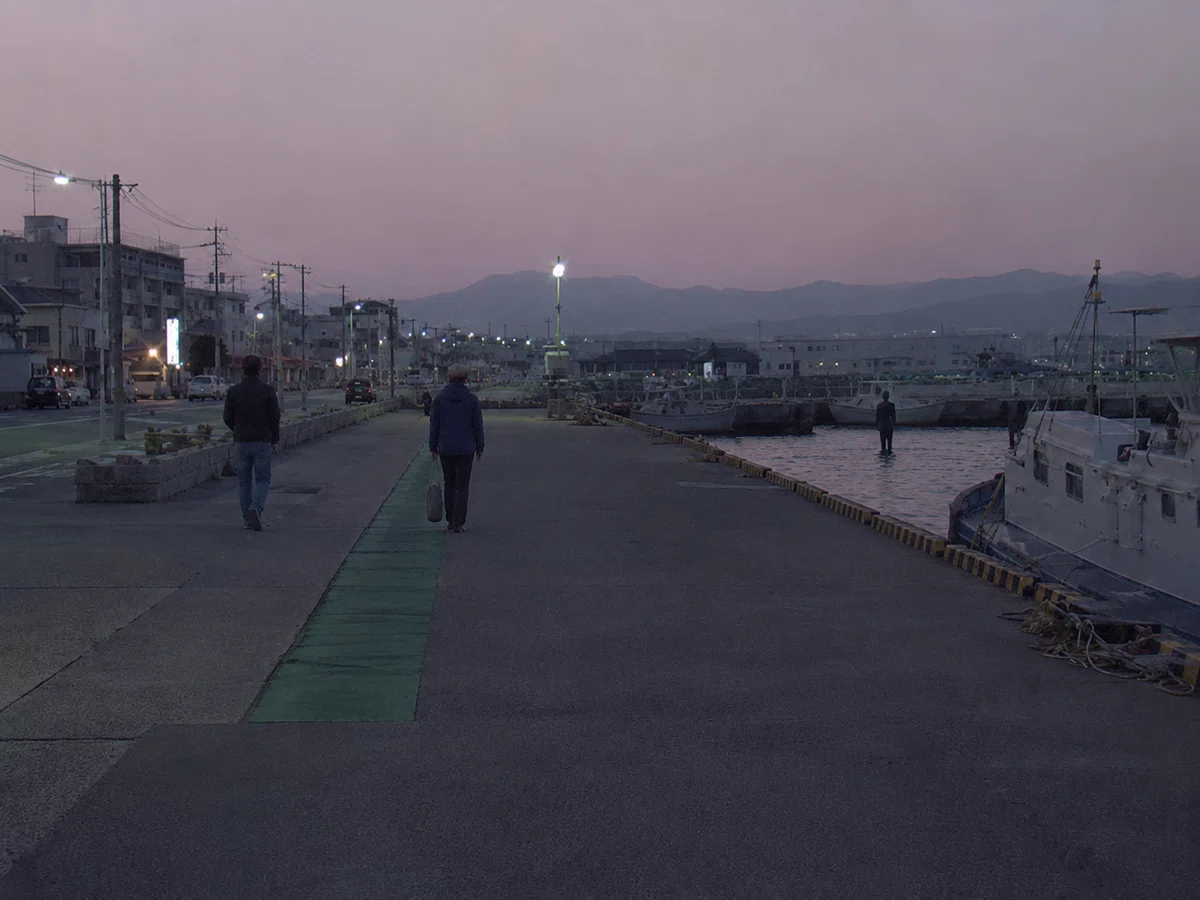 夕暮れから夜にかけての港沿いの道路を撮影した記録写真。海沿いの歩道付近には一般の歩行者が二、三人見え、日常的な港の風景に見える。画面の海側、水際または浅い海の中に、細長い体格で肩幅の広い黒い背広姿の人物のようなものが立っている。周囲の歩行者は特に反応しておらず、写真全体も港沿いの道を撮影したものに見える。対象は主題として撮られたのではなく、画面の端に偶然写り込んだように見える。