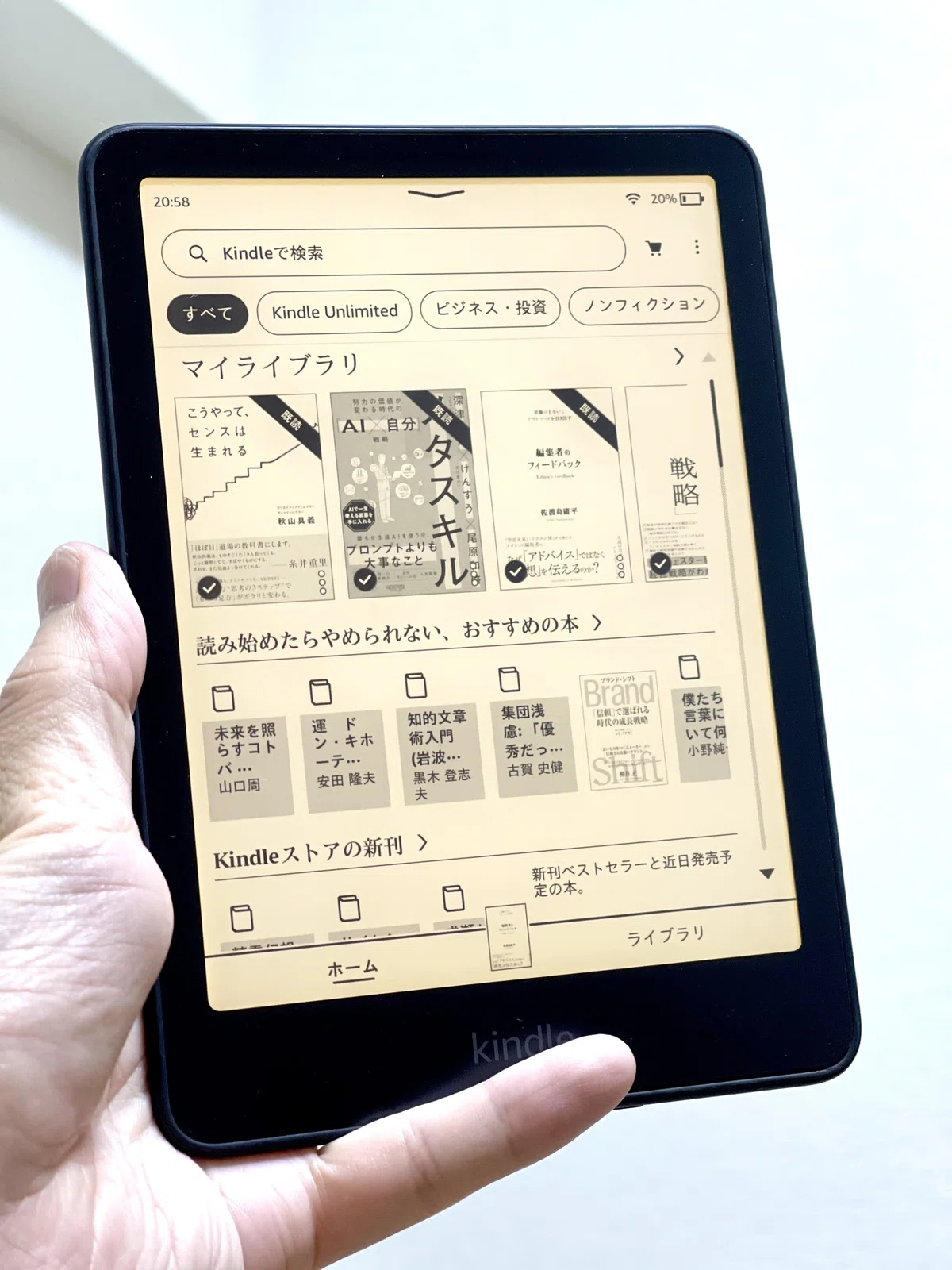 片手で持ったKindle電子書籍リーダーの画面に、マイライブラリの書籍カバー一覧や『読み始めたらやめられない、おすすめの本』などのストア推奨本が表示されている様子