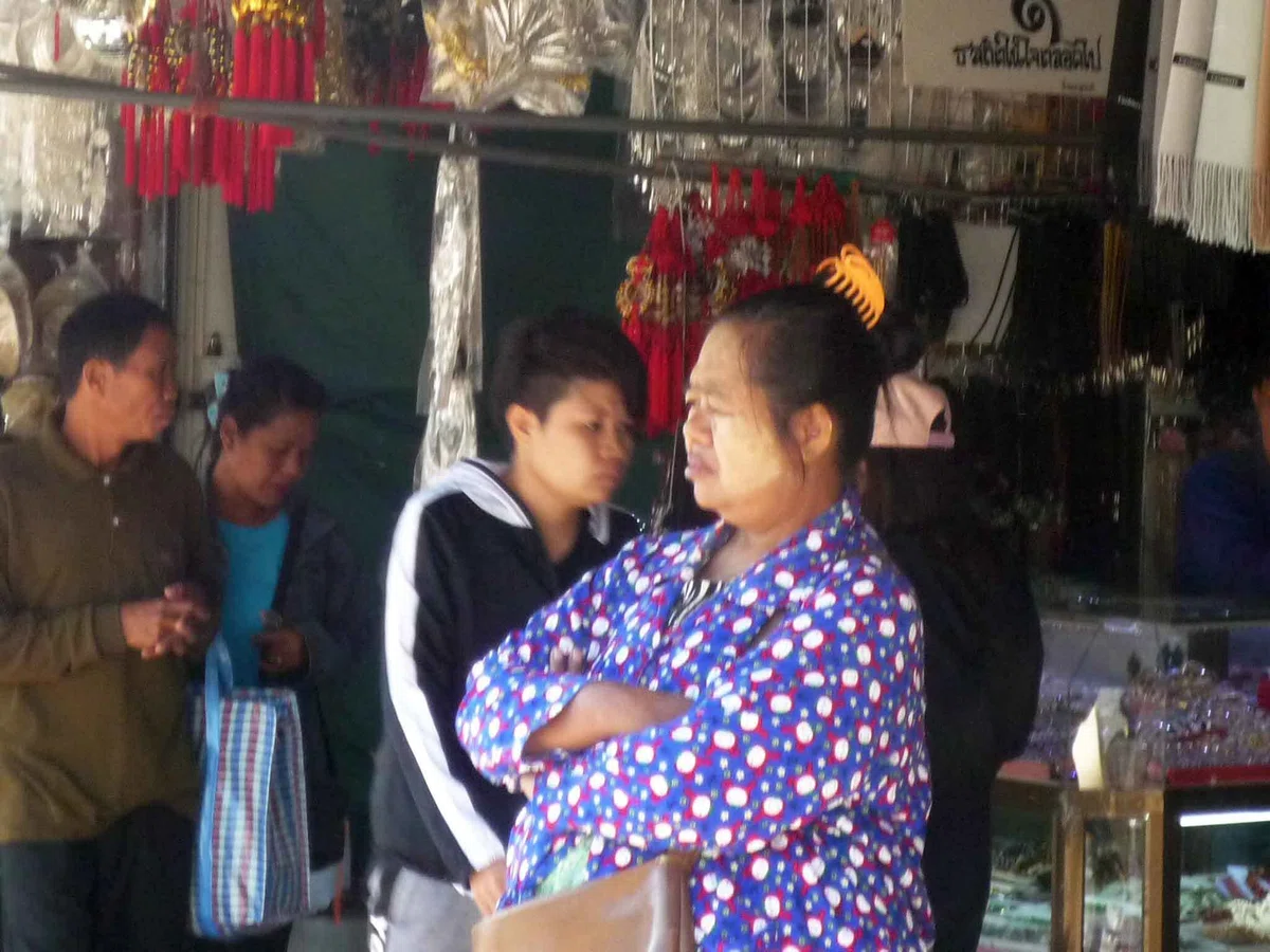 店先で頬に白っぽい化粧をした女性が腕を組み、後ろに数人が立っている