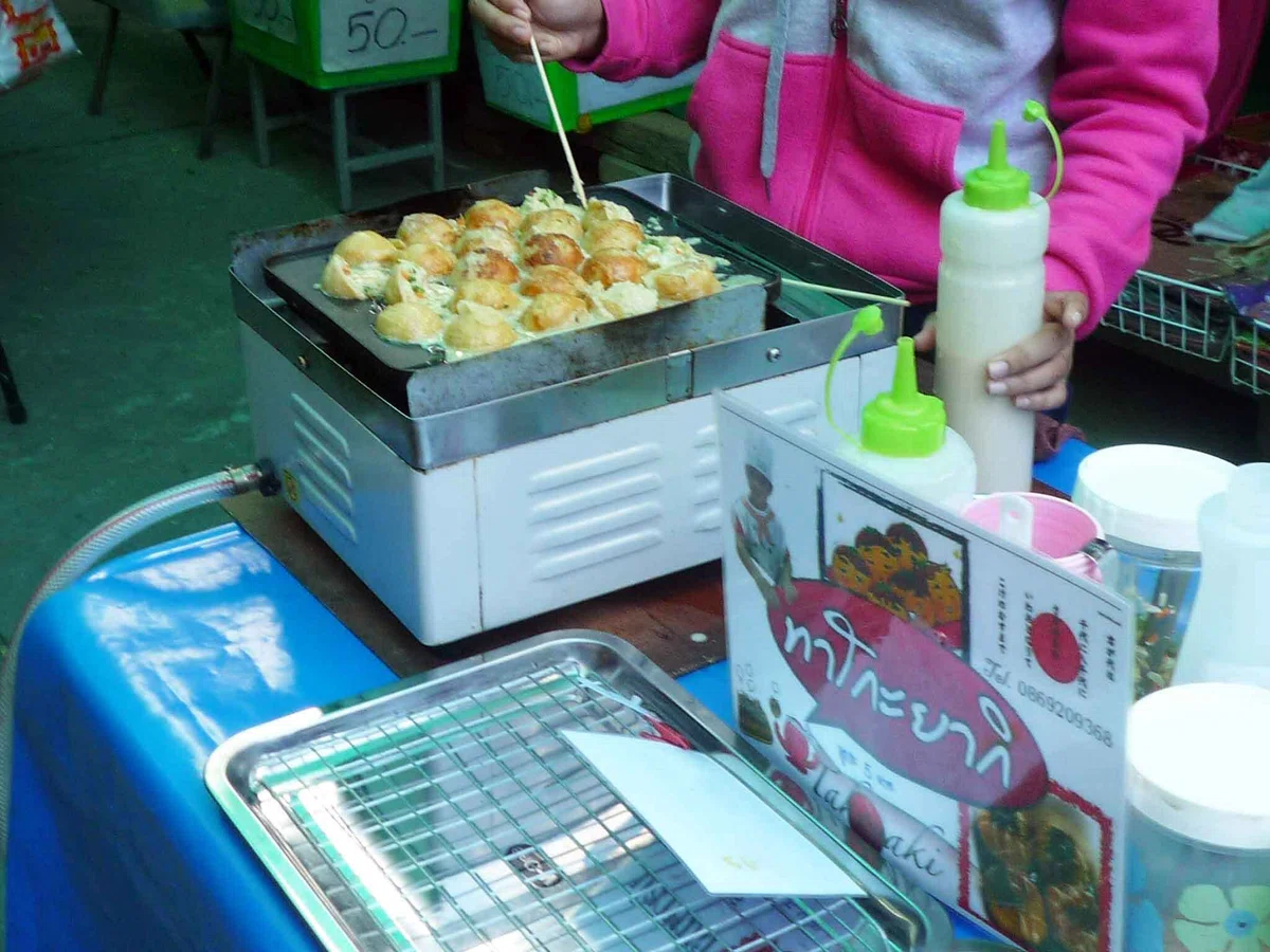 日本国旗をあしらった看板の横で、店員が鉄板のたこ焼きを竹串で焼いている
