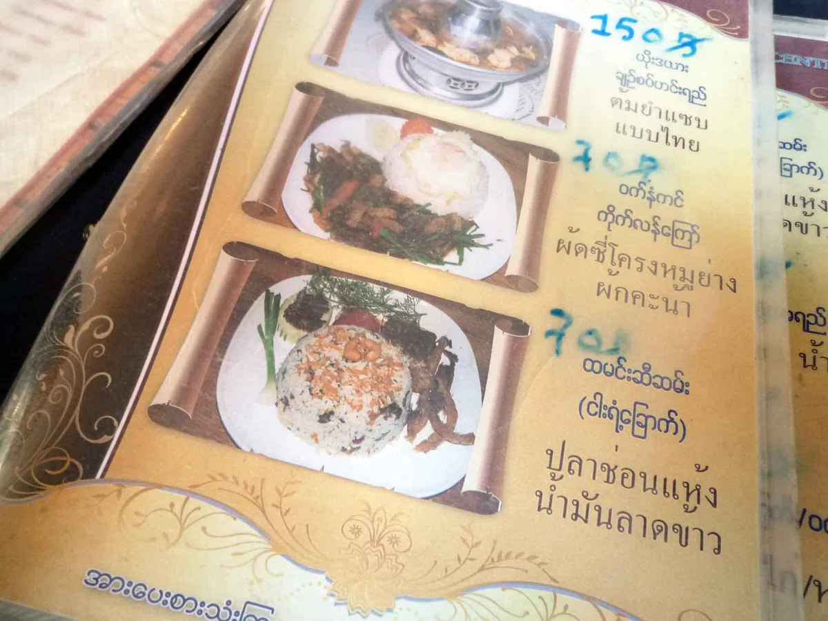 料理写真の横にミャンマー文字とタイ文字、手書きの価格が記されたメニュー