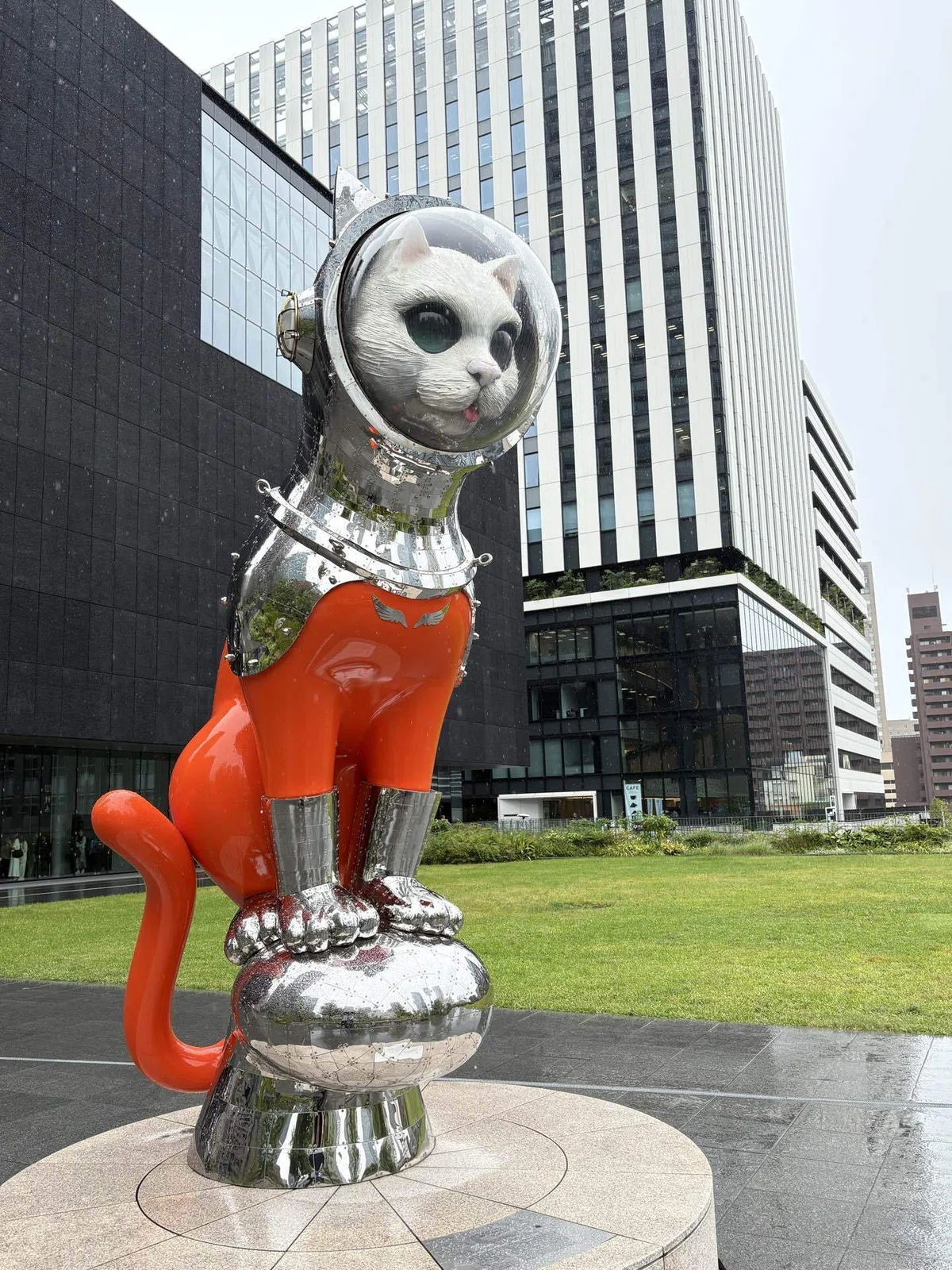 大阪中之島美術館の屋外に設置された、宇宙服を着た猫の大きな彫刻。オレンジ色の体と銀色の宇宙服が印象的で、背景には美術館周辺の建物と芝生が広がっている。