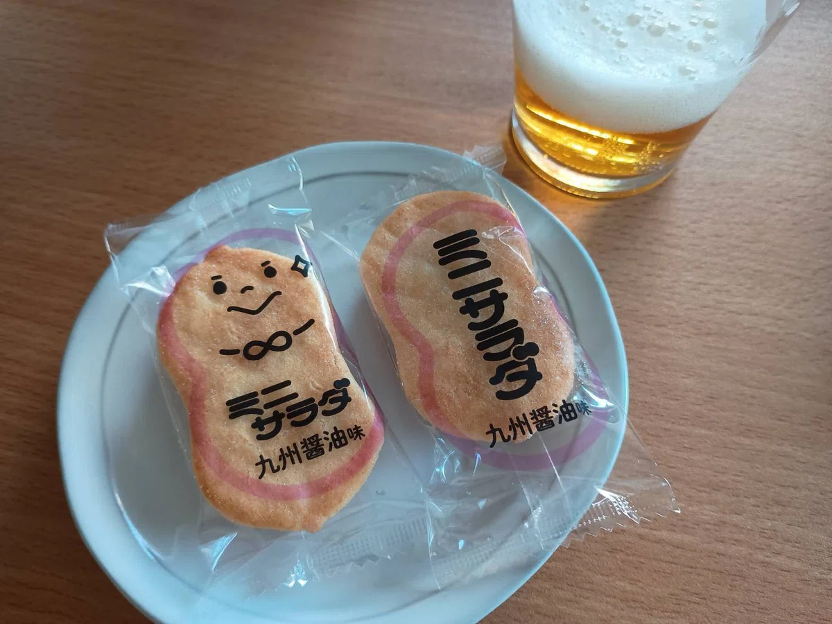 お煎餅のミニサラダの九州醤油味二つで、片方に顔のプリントがある、ビール
