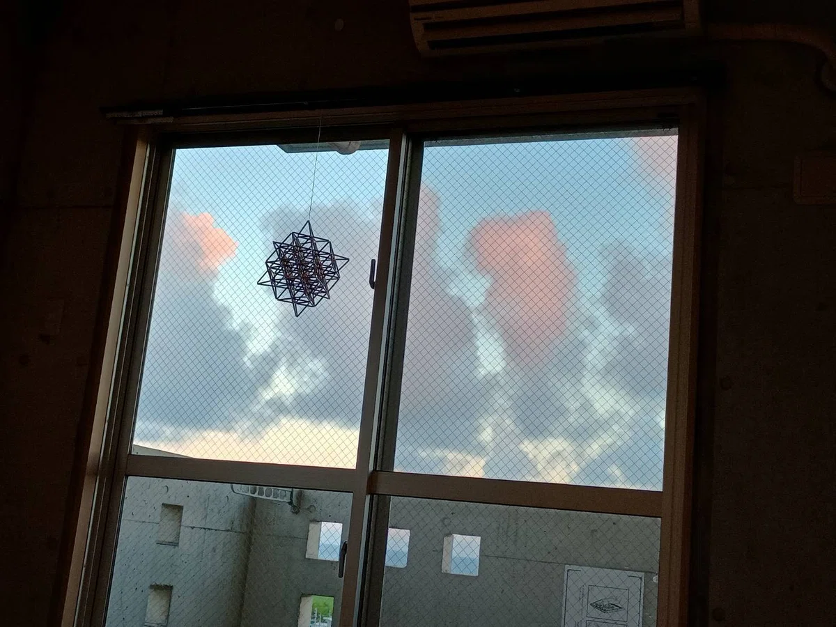 部屋からベランダ側の窓を撮ったもので、窓の外にはピンク色とグレーの雲がモコモコと立っている様子があり、ベランダの格子の穴からは海と緑色の植物がのぞく、窓辺にはストローで作った64テトラヒドロンが飾ってある