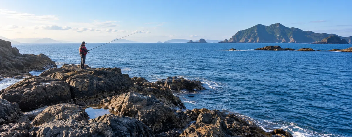 救命胴衣を着けた釣り人が、穏やかな磯から海へ竿を出す様子