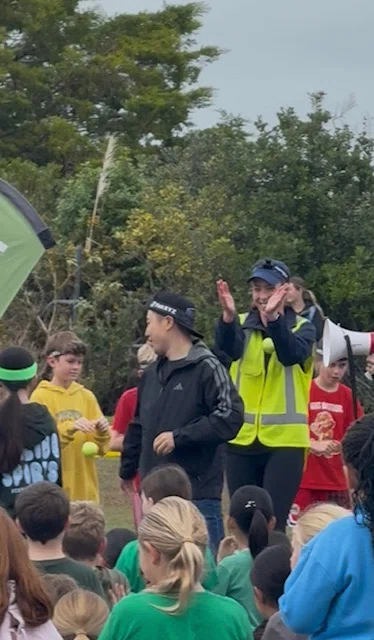 A woman in a high-visibility jacket applauds during an outdoor school event with children gathered around.