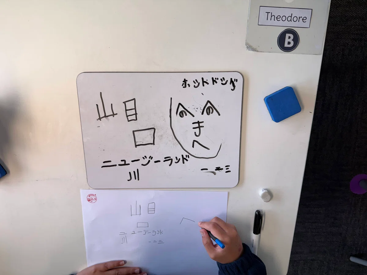 A student copies Japanese characters and a henohenomoheji face drawing from a small whiteboard onto paper.