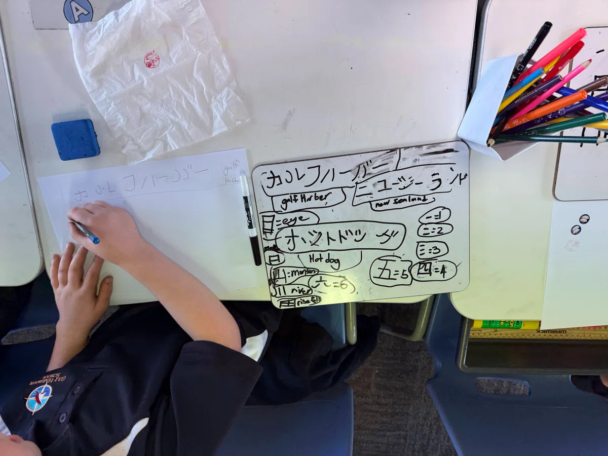 A student writing Japanese characters on paper beside a reference sheet on a classroom desk.