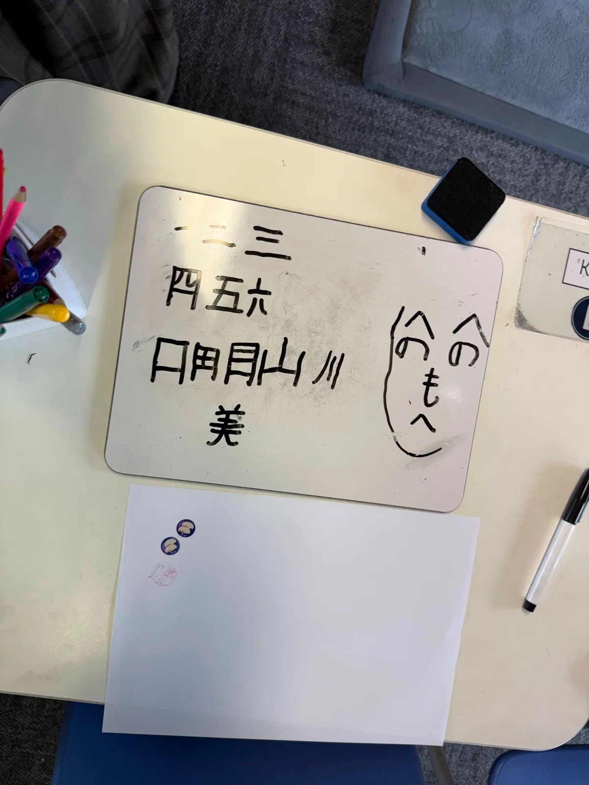A sheet of paper with handwritten Japanese kanji, including the character 美, and a henohenomoheji face drawing.