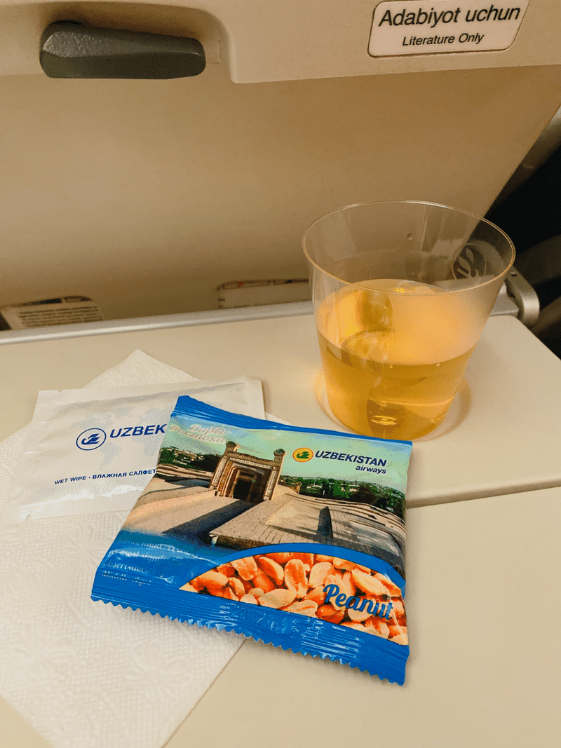 ウズベキスタン航空のロゴが書かれたピーナッツのお菓子の袋と、お茶が入った透明のコップ。どちらも飛行機の座席テーブルの上に乗っている