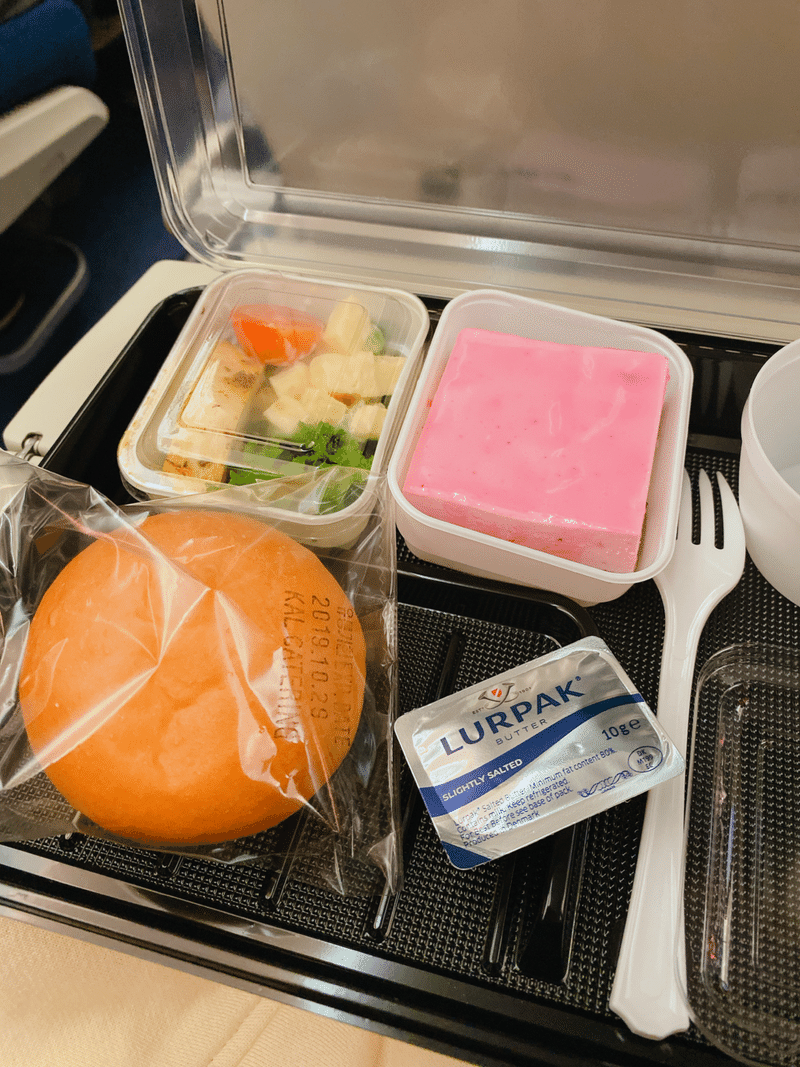 ウズベキスタン航空の機内食。丸い形のパンとバター、ピンク色をしたババロアのようなお菓子、容器に入った惣菜がプレートの上にカトラリーと合わせて載せられている