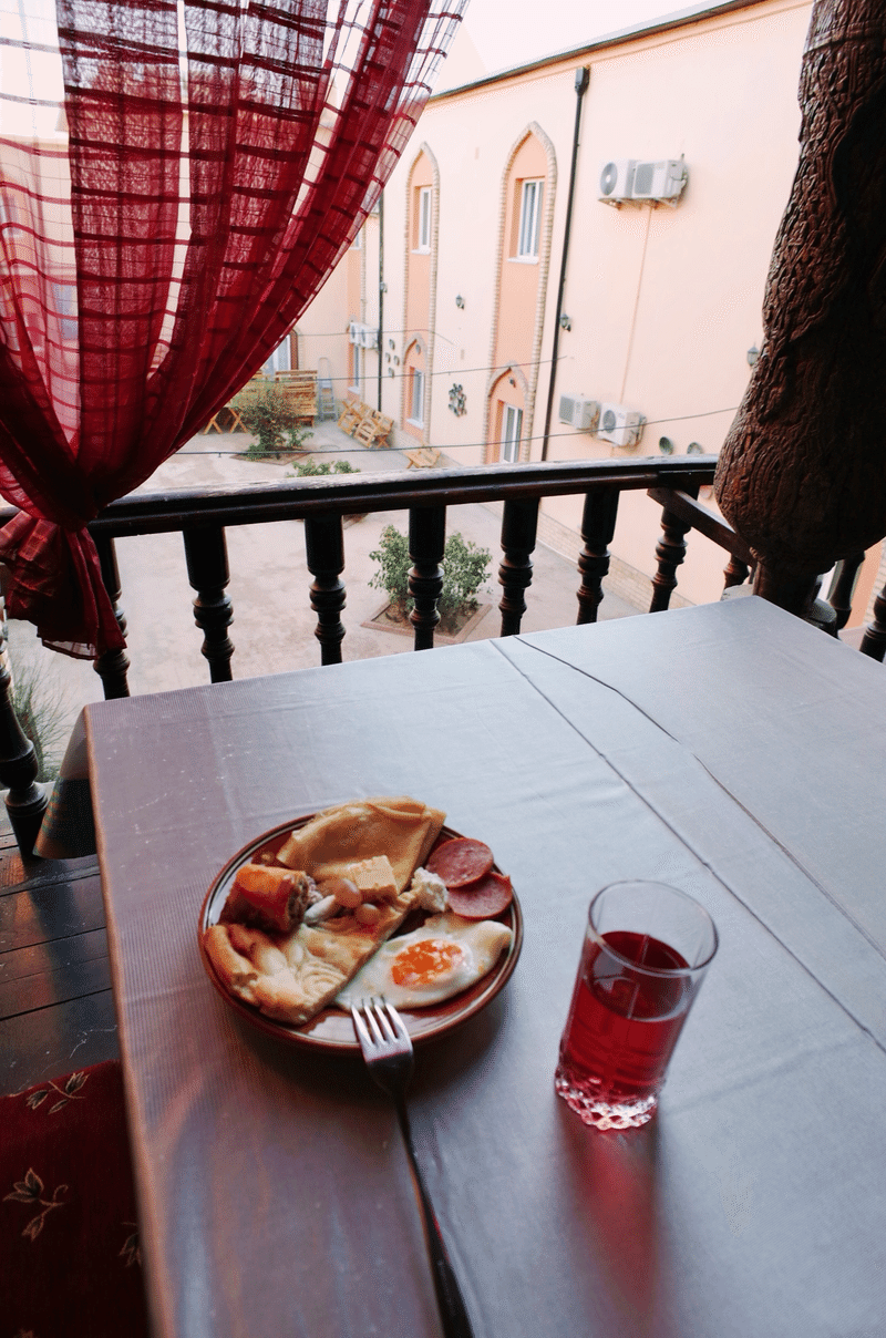 中庭に面したテラス席のテーブルに、プレートひとつとジュースが置かれている。プレートには目玉焼きやハム、薄い形状のパンなどが乗っている