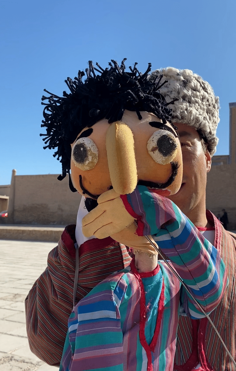 ウズベキスタンの男性を模した人形を操り、人形の左手で人形の口をおさえて驚いた表情をつくっている様子