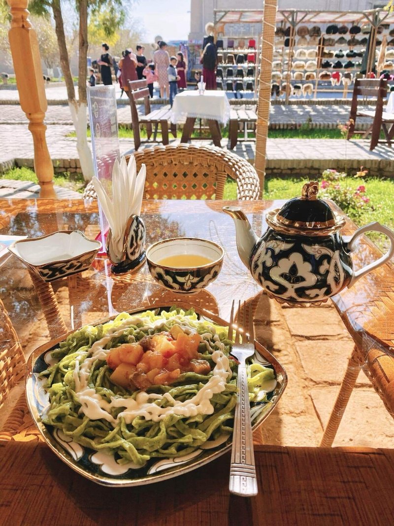 背景は人でにぎわう通り。テーブルの上には緑色の麺の料理が乗ったお皿とフォーク、お茶のポットとカップが置かれている