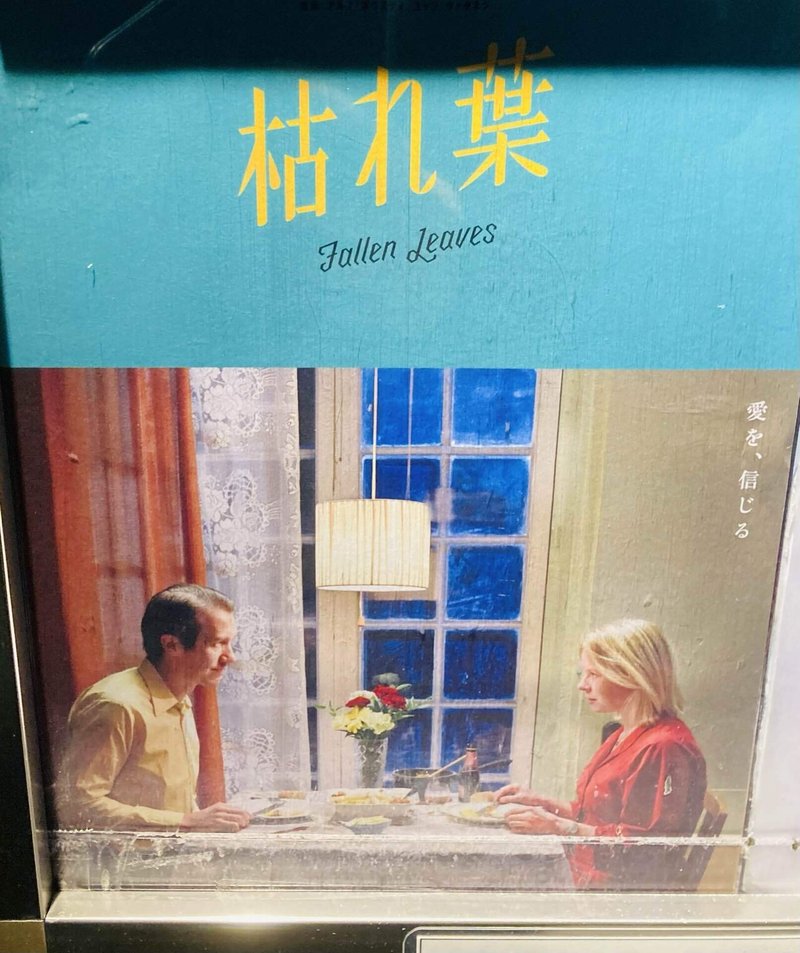 『枯れ葉』のポスター。上部背景はターコイズ色、ロゴは黄色で書かれている。下部は映画のワンシーン。主人公二人が食事を共にしている。