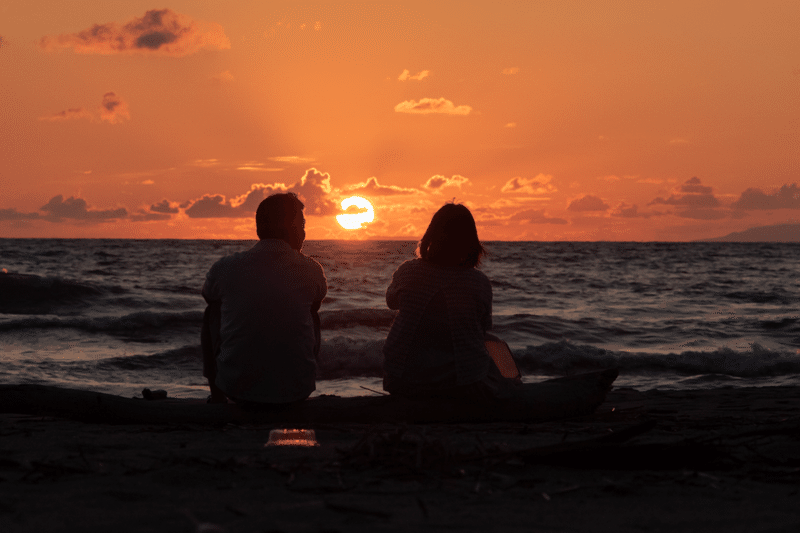 海辺にて、夕日の沈みかけた赤く染まる夕空を、味のある距離感で座って語らう熟年夫婦のシルエットが素敵。