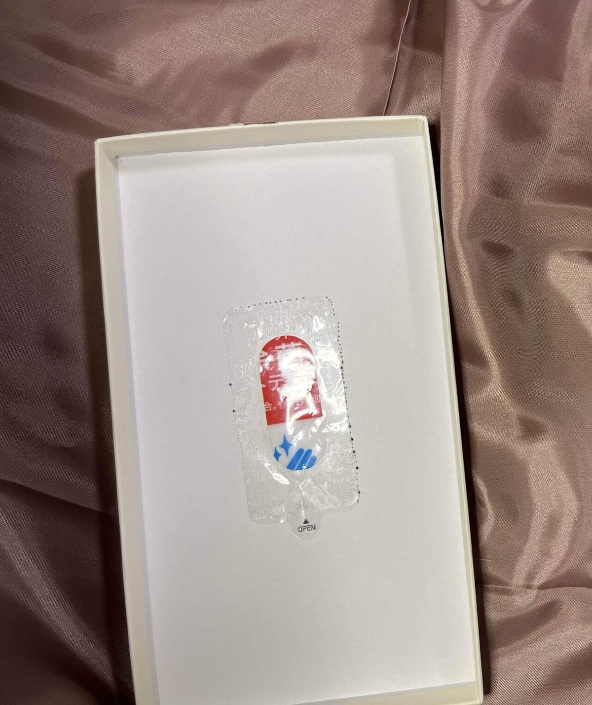 写真には、白い箱の中に透明なプラスチックの包装が入っています。包装の中には赤と青のデザインが見えます。箱は柔らかいピンク色の布の上に置かれています。包装には「OPEN」と書かれたタブが付いています。