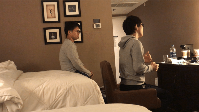 ホテルで発表練習している様子。小林が身振り手振りを使って話し、齋藤さんが後ろでじっと見守っている。