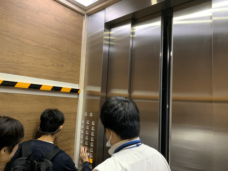 写真：人荷用エレベーターに乗りスイッチを操作する杉崎くんと社員