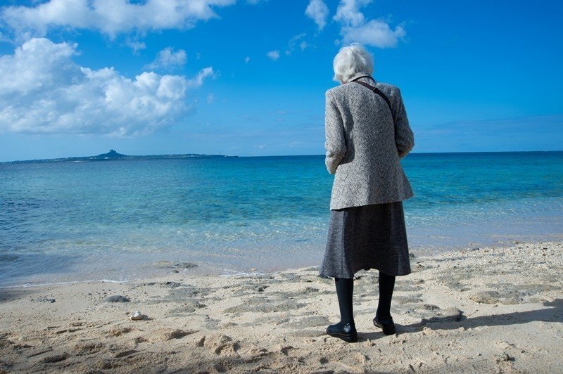 オキナワへいこう場面写真　青い海、砂浜に立つ老婆の後ろ姿