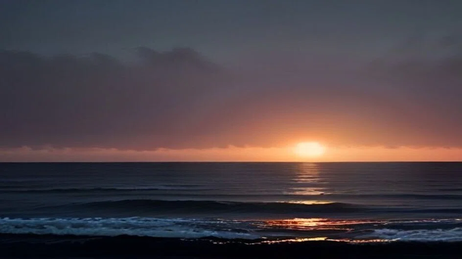 夕暮れの海を低い位置から見たところ。厚い雲の下に橙色の帯が広がり、沈みかけた光が水面に長い道をつくっている。手前では白い波が崩れかけている