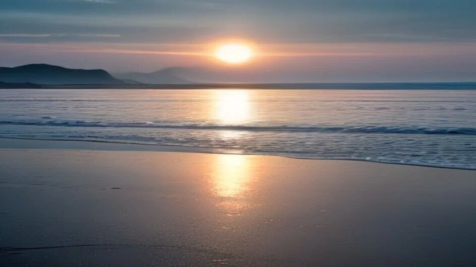 夕方の浜辺。低くなった日が海に落ちかけていて、その光が濡れた砂の上へまっすぐ伸びている。奥には霞んだ岬が見える。人はいない