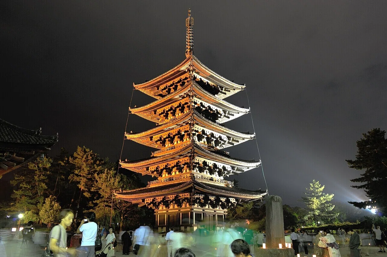 夜の木々に囲まれ、ライトアップされた五重塔。訪れる人影がぼっと浮かぶ、奈良の夜の情景です。