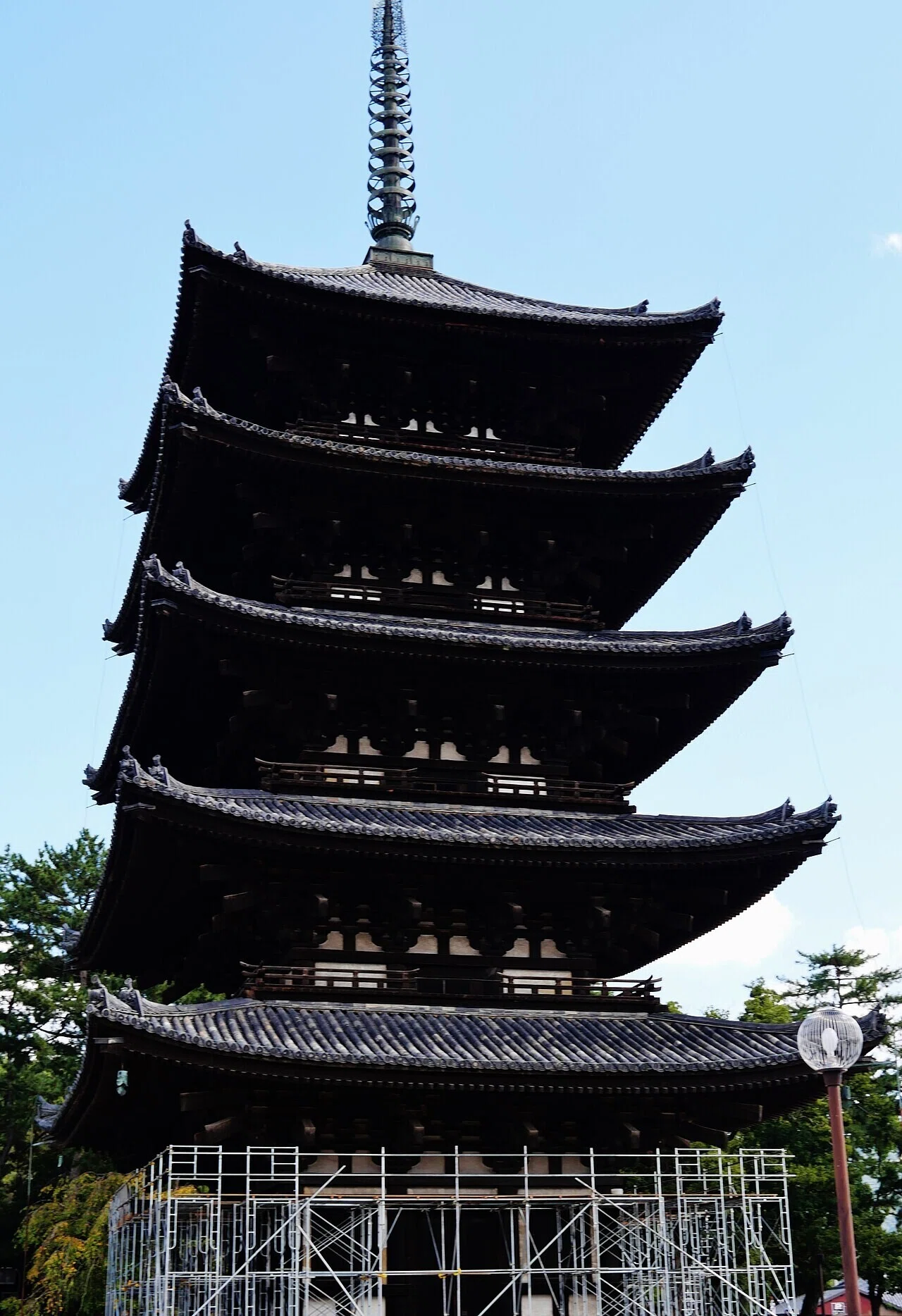 青空を背景にした瓦屋根の五重塔。基壇部に足場が組まれ、修理保存の作業が行われている様子が分かります。