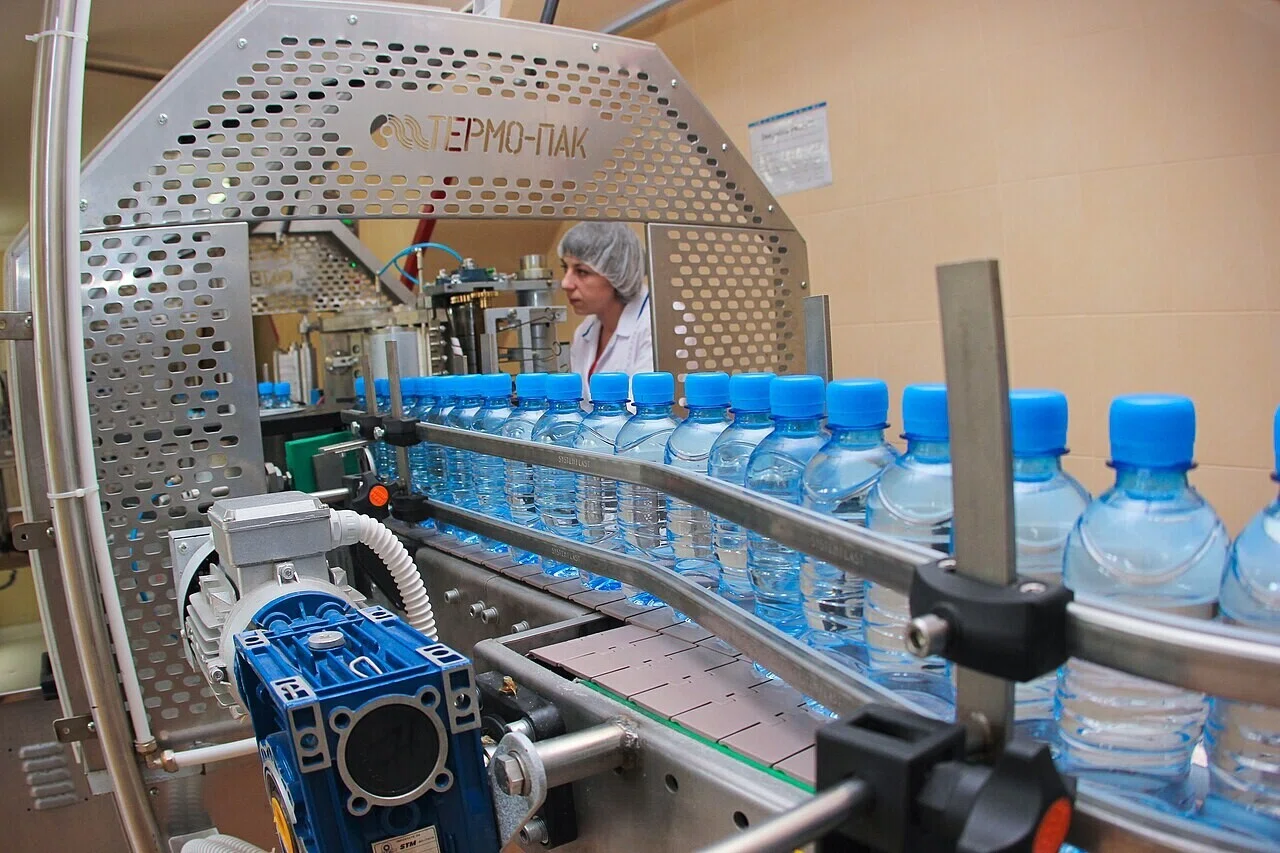 工場のベルトコンベアを流れるペットボトルと、背後で作業する作業員の姿。大量生産されるモノもまた、数えられ管理される対象だった。次の節はその起点にある帳簿をたどる。