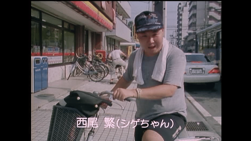 場面写真　野球帽で自転車にまたがったシゲちゃん（西尾繁）